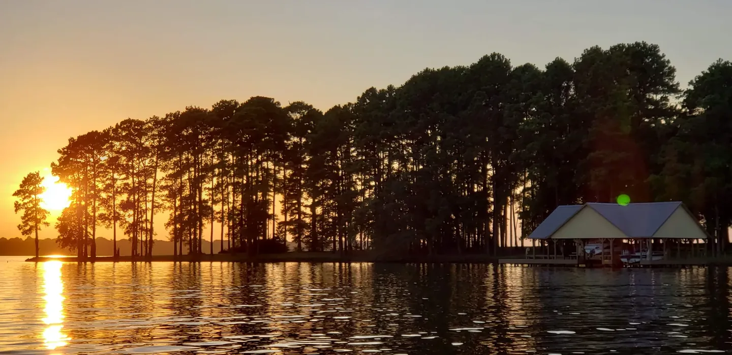 A covered two-slip boathouse with a metal roof sits at the end of a pine-lined point on Lake Palestine at sunset, with a boat moored under the roof and calm open water in front of it.