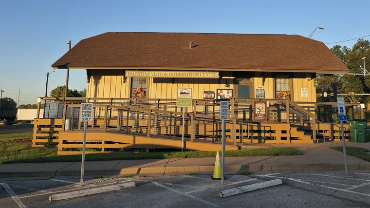 The Palestine Visitor Information Center, a tan building with a brown roof, a ramp and a wraparound deck, seen from its parking lot.